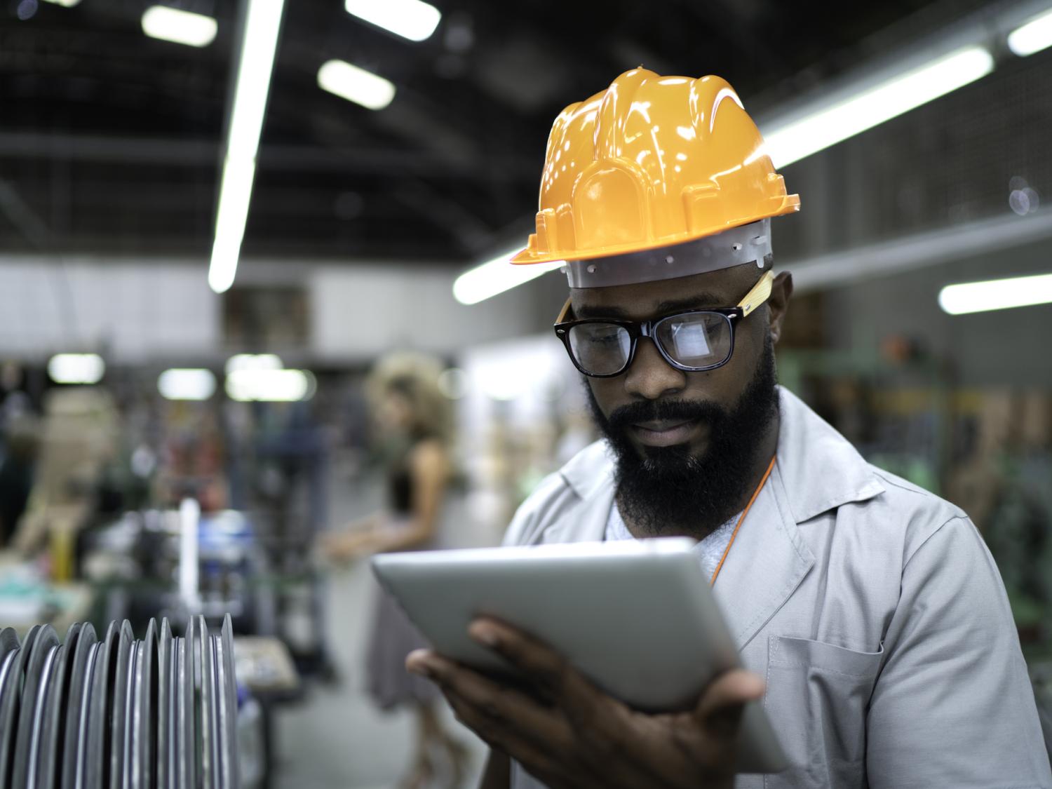 Man in a yellow hardhat looks at a tablet while standing in a manufacturing facility