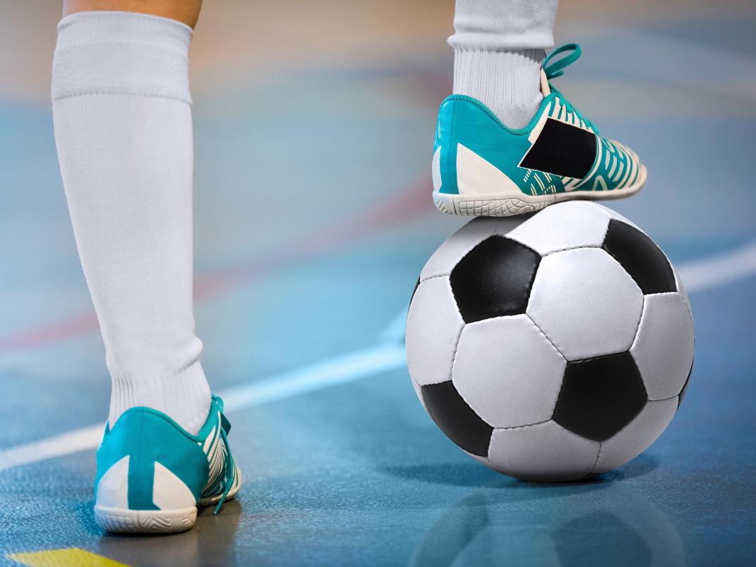 Close-up of an athlete’s legs as they stand on a gym floor, the right foot resting on a soccer ball.