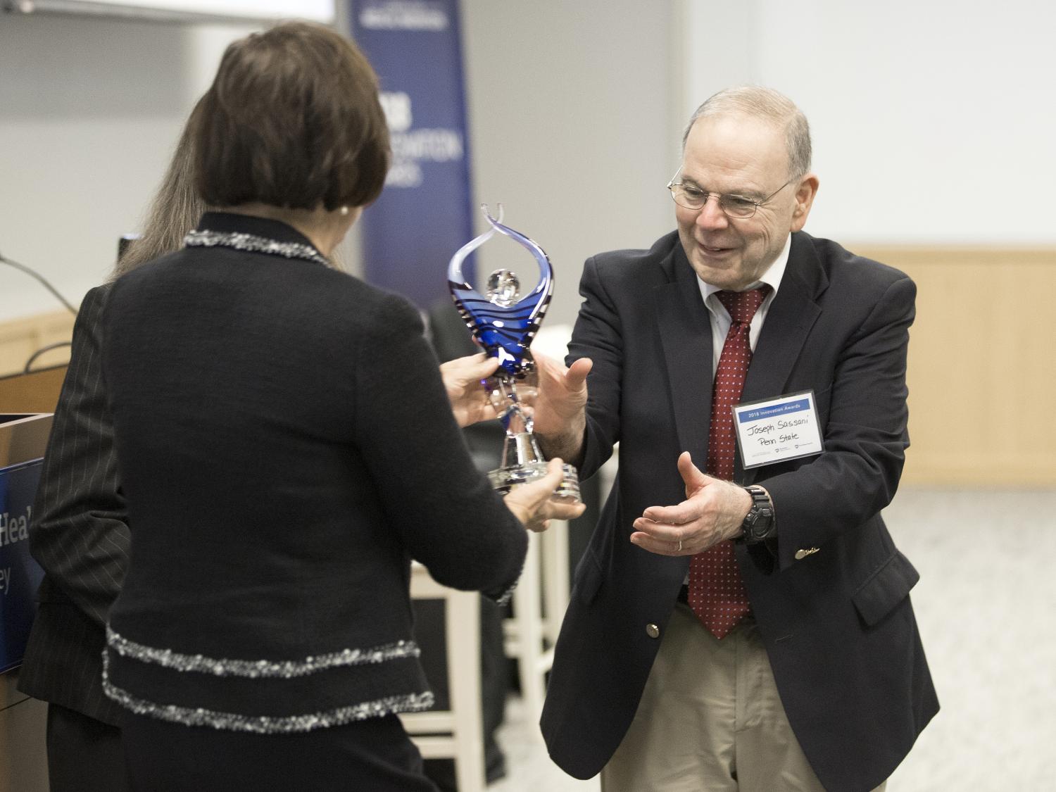 Two women and a man, all in semi-formal attire, reach out and jointly hold a small glass statue, being given as an award.