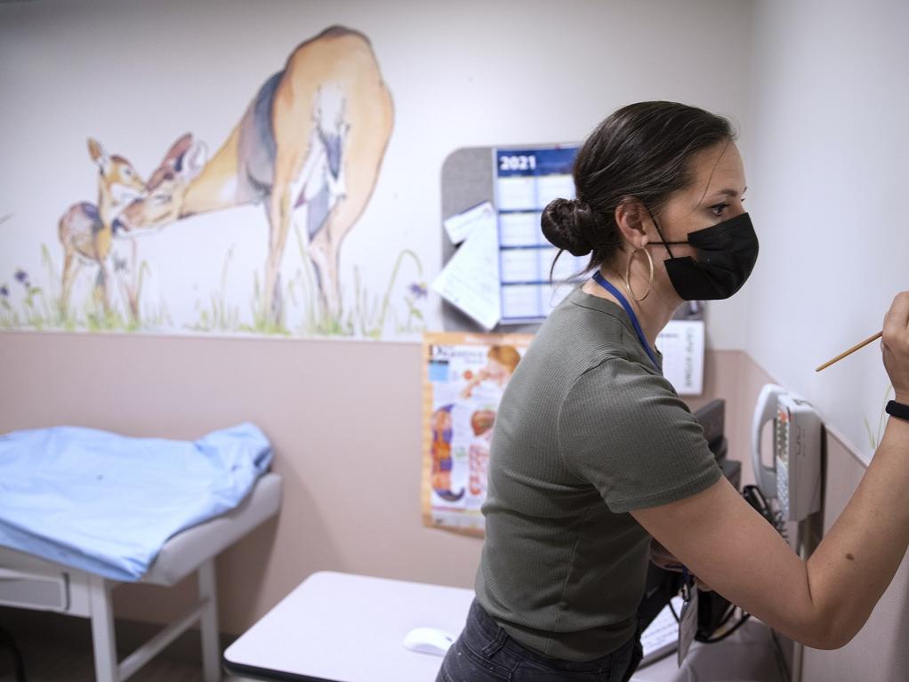 A woman in a surgical mask applies a paintbrush to a hospital room.