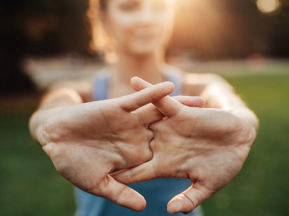 A close-up of a woman’s hands with fingers interlaced with palms facing outward. The woman is out of focus, as is the green background, which is grass.