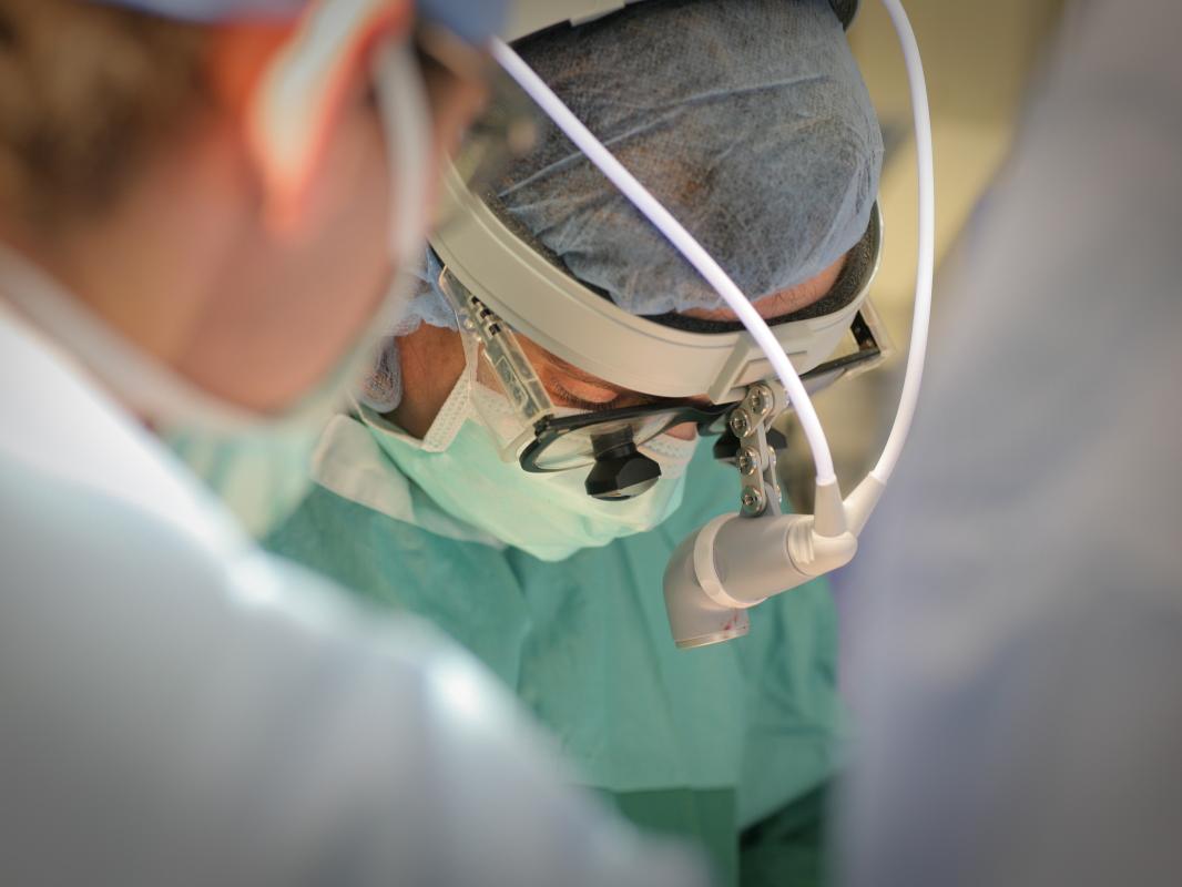 Dr. Zakiyah Kadry, wearing a hair net, surgical headgear, a face mask and a surgical gown, looks down at the operating table. Another staff member is near the camera, out of focus.