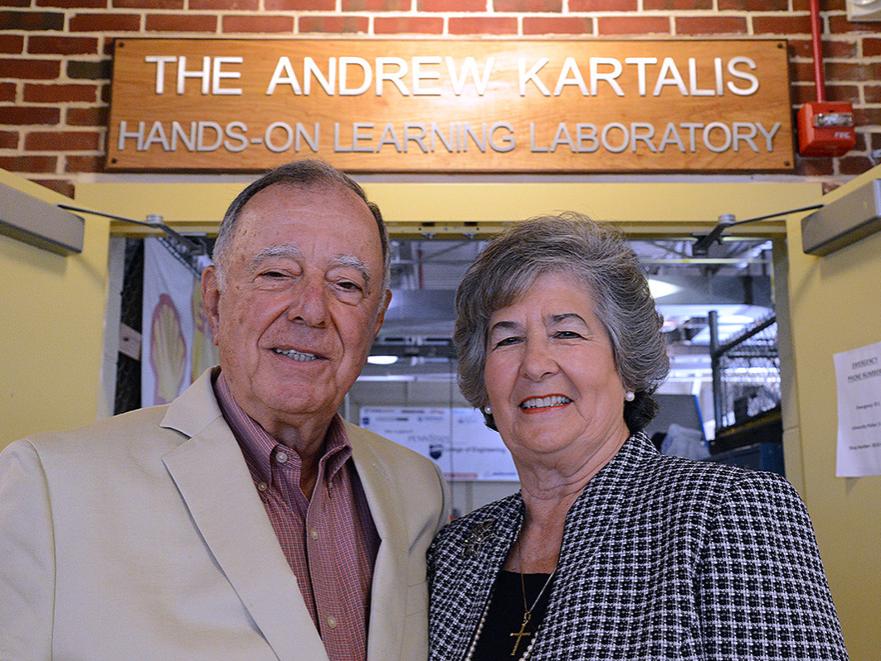 A man and woman smile at the camera. The background, a sign reading, "The Andrew Kartalis Hands-On Learning Laboratory," can be seen. 