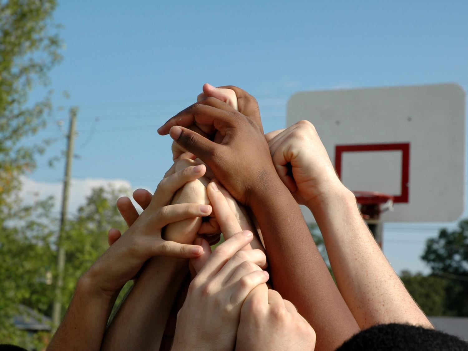 Close-up of several children extending their arms into the air to form a pyramid. In the background are trees, power lines and a basketball hoop.