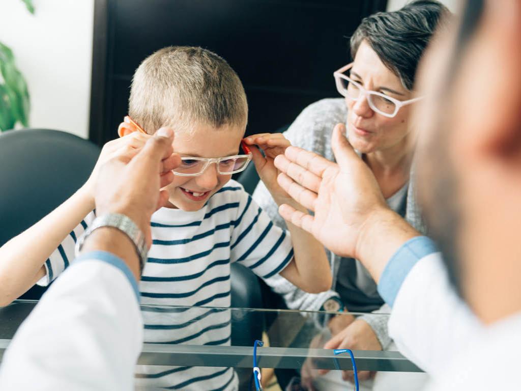 A young child wears glasses, holding onto the frames with their fingers. A woman is seated beside the child, and an individual in a white coat is seated next to the camera, with arms extended toward the child’s glasses.