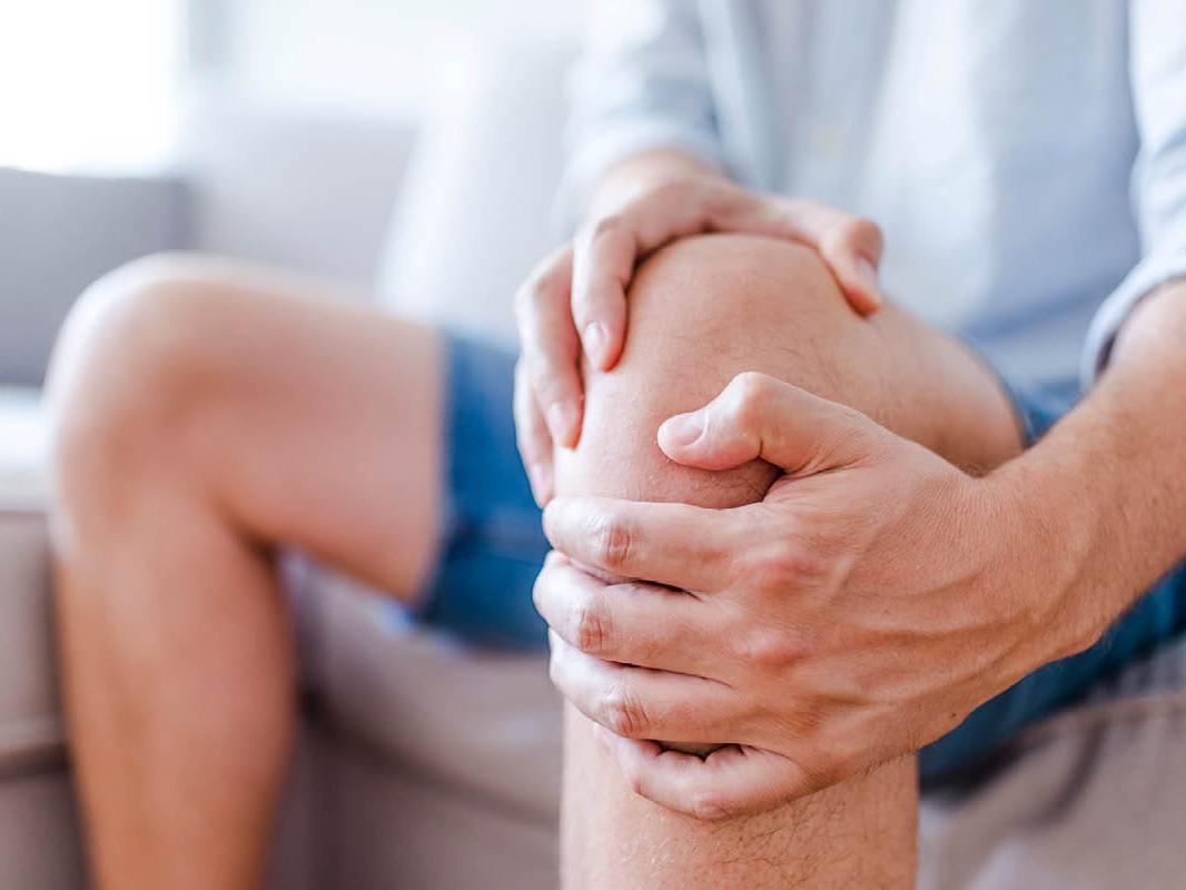 Close-up shot of a man, seated on a couch, holding his left knee with both hands.