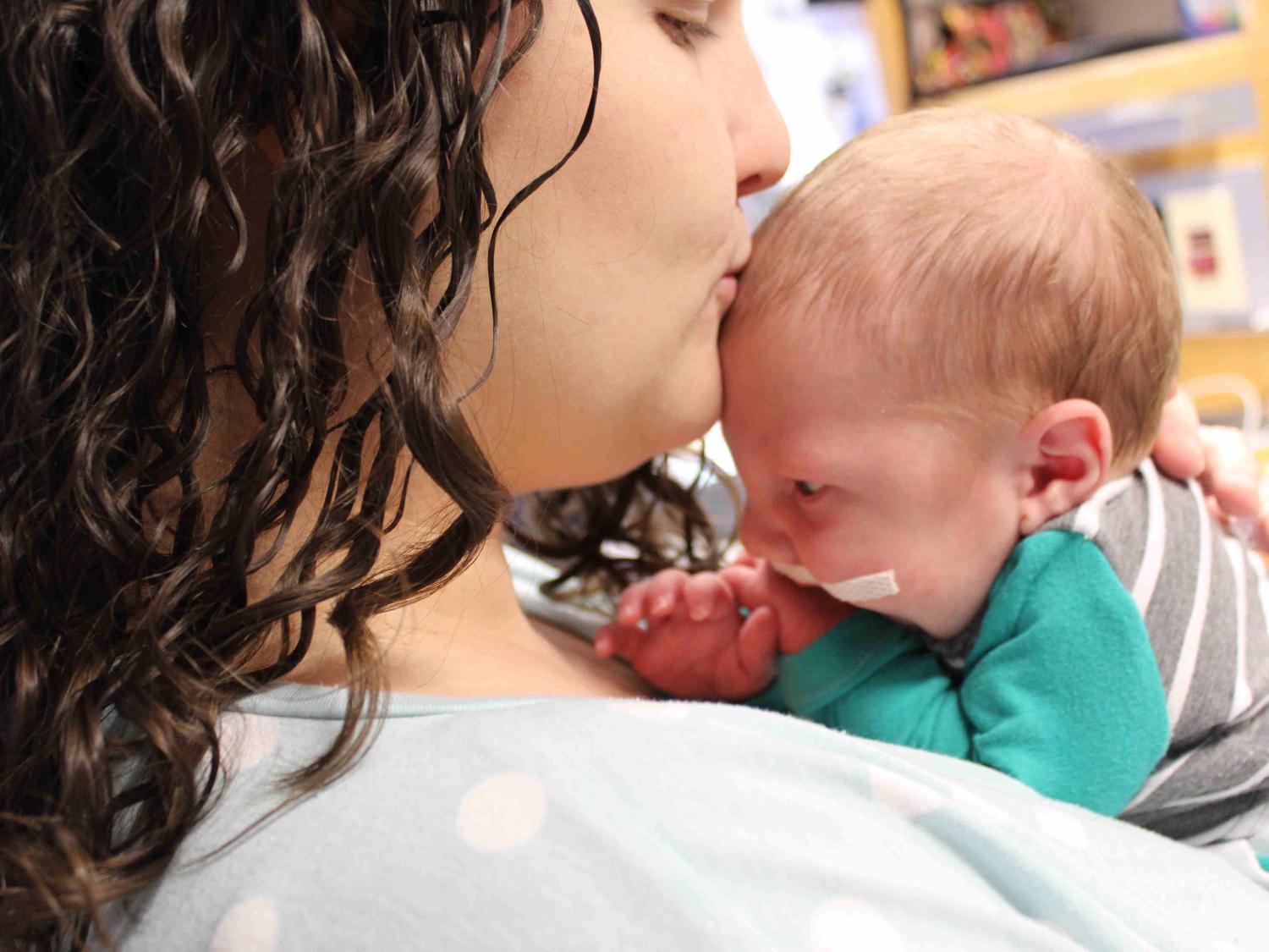 Natasha Himes kisses her newborn baby boy as she holds him on her chest. He has surgical tape on his face and his wearing a striped shirt. Natasha has long curly hair and is wearing a cotton top.