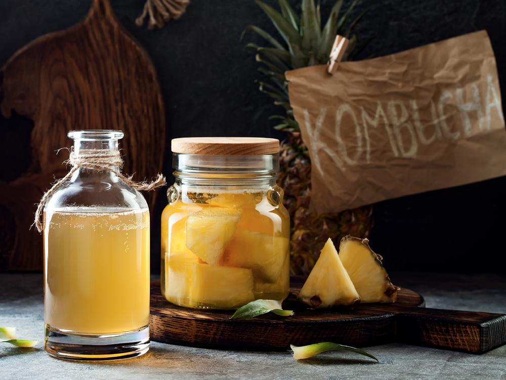 In the foreground are two glass jars with yellow liquid, one containing pieces of pineapple. In the background is a pineapple and a sign reading “KOMBUCHA.”