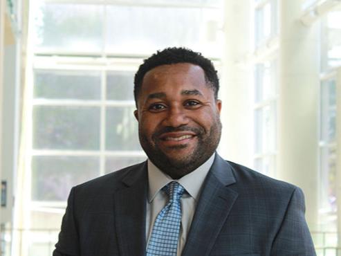A headshot of a man smiling at the camera and wearing a gray suit jacket, white shirt and light blue tie. 