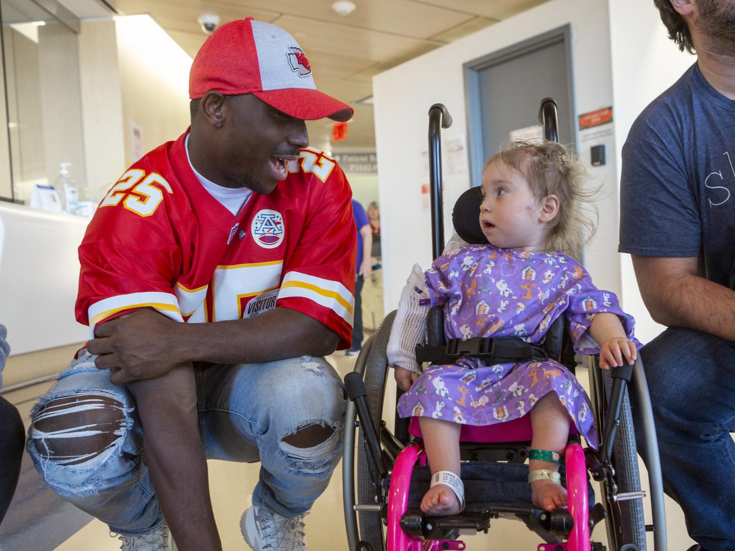 LeSean McCoy, wearing a Kansas City Chiefs uniform and hat, kneels down to talk with a young girl in a wheelchair. She looks back at him.