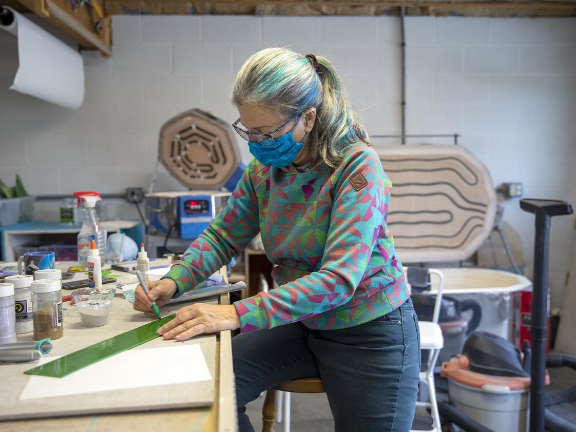 Linda Billet perches on a stool in her workshop and draws with a marker on a strip of glass. She wears glasses and a face mask and has her hair in a ponytail streaked with colors. Art supplies surround her on the counter where she works.