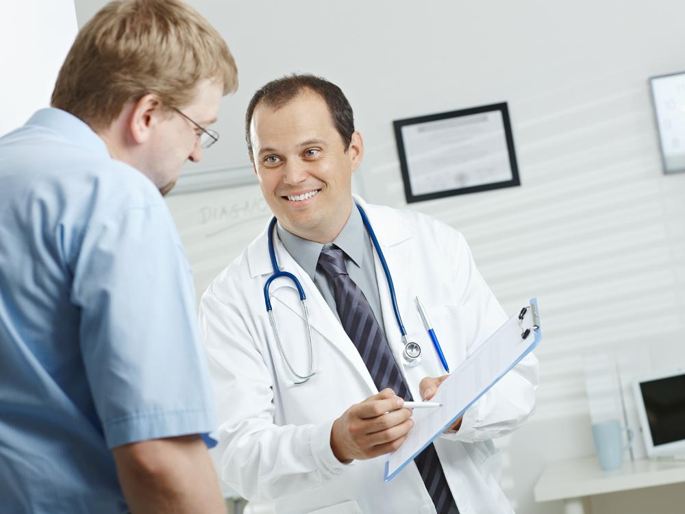 A male physician wearing a white coat and a stethoscope around his neck holds a clipboard with his left hand, using a pen in his right hand to point to the clipboard as a male patient looks down at it.