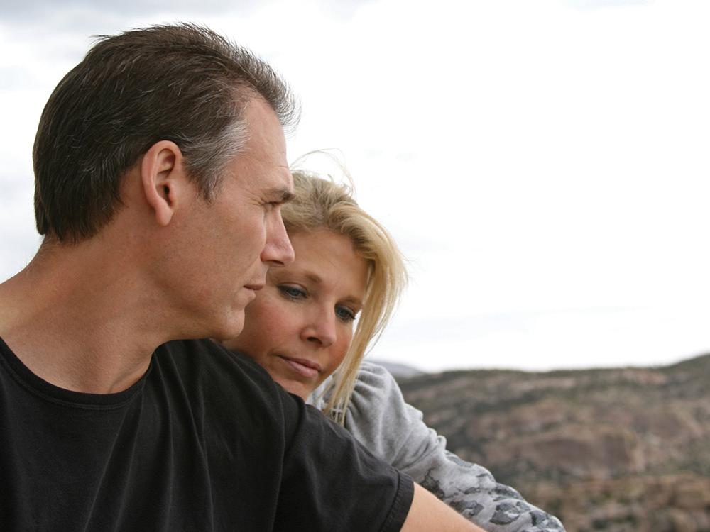 A man facing the camera looks over his left shoulder. A woman has her head resting on the upper part of his left arm. A hill is in the background, out of focus.