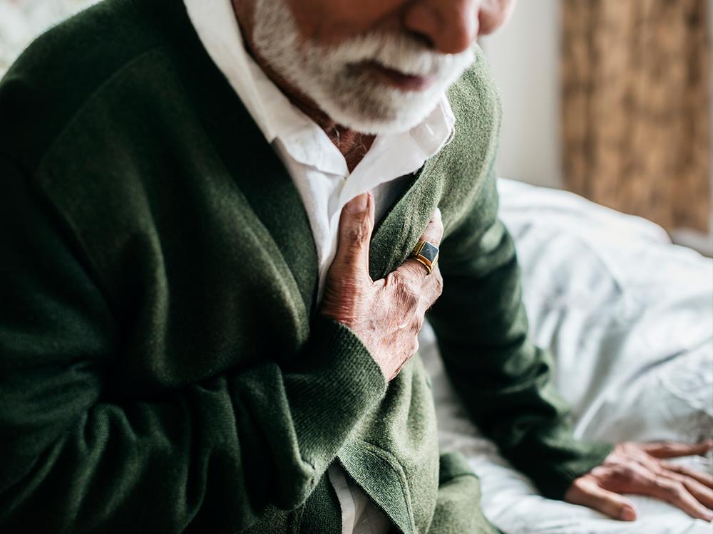 A close-up of a seated man, leaning slightly forward and clutching at his chest with his right hand.