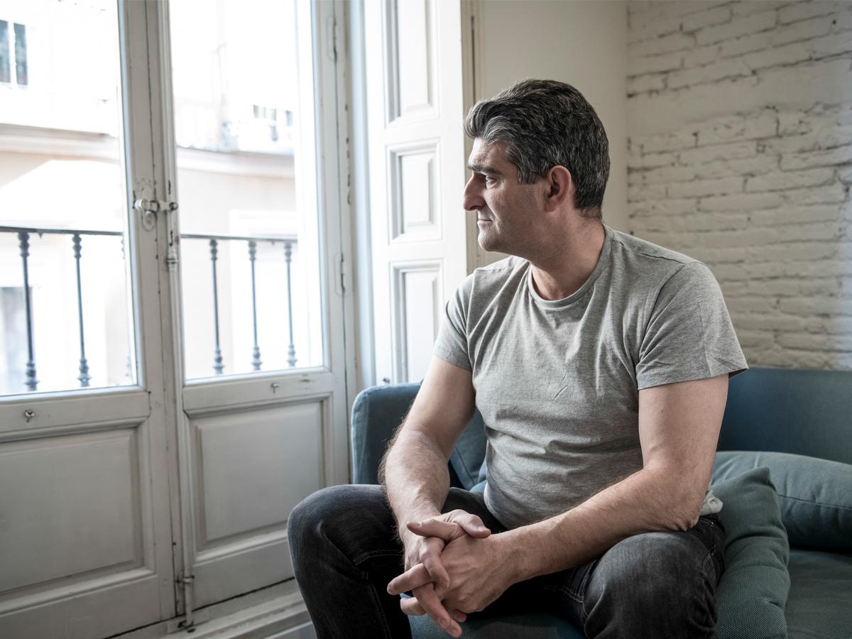 A middle-aged man sits on a couch, looking out the window of the room he’s in.