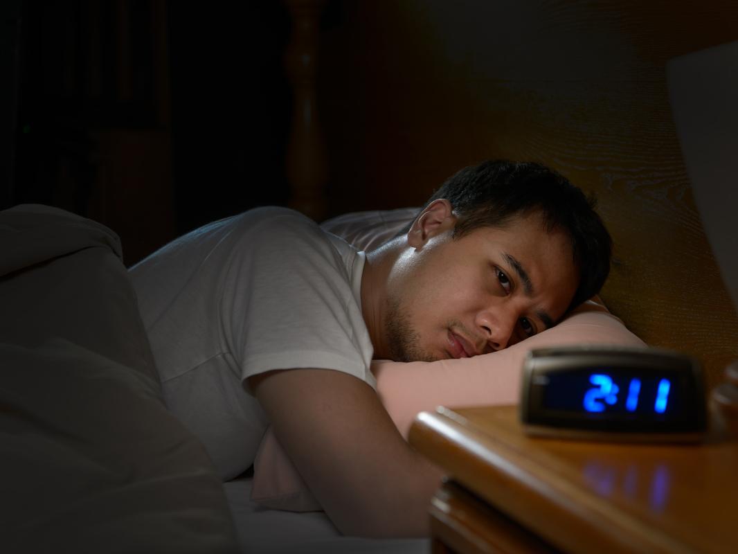 A man wearing a white shirt looks at an alarm clock while experiencing trouble sleeping