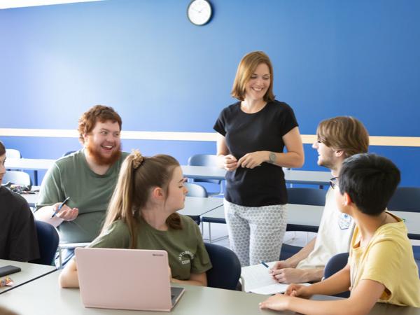 A female professor in a black t-shirt stands and interacts with a group of seated students
