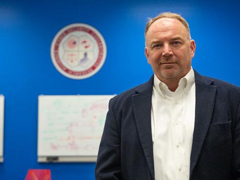 A man in a white button down and navy suit jacket poses in front of a blue wall and a whiteboard. 