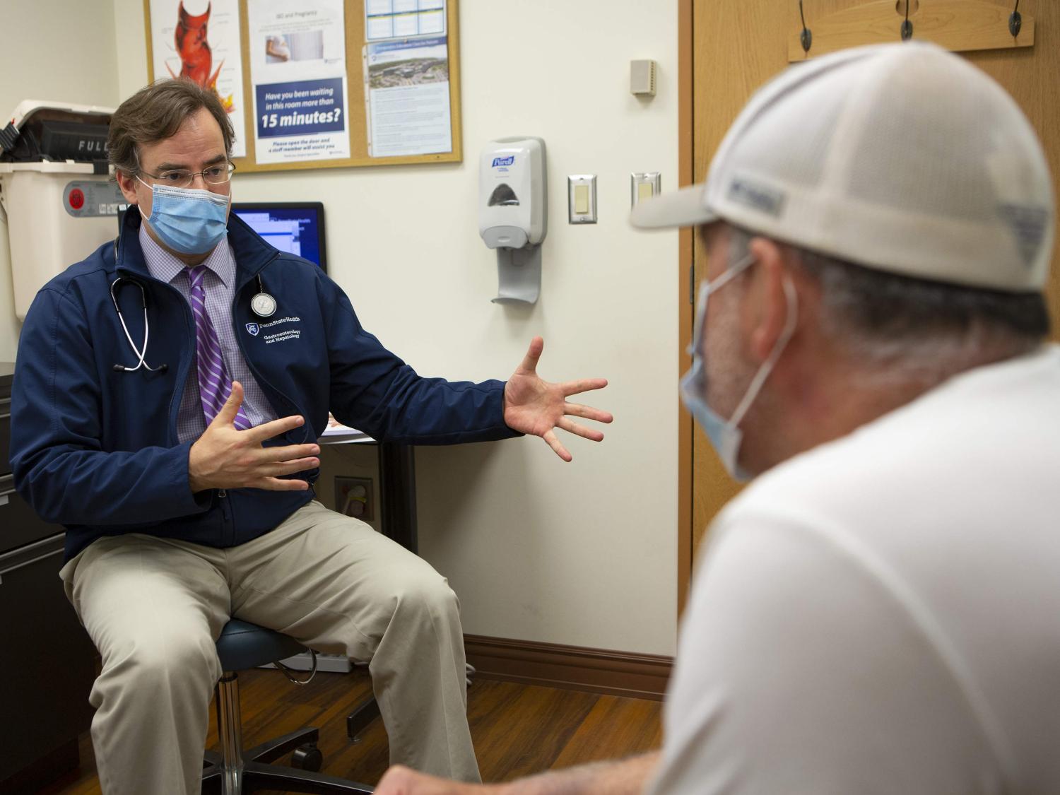 A male physician wearing a tie, Penn State Health coat, face mask and stethoscope, gestures with his hands while talking with a man seated across from him in a clinic room.