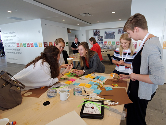 Five people standing around and leaning on a table covered with Post-its, pipe cleaners, construction paper and cups. 