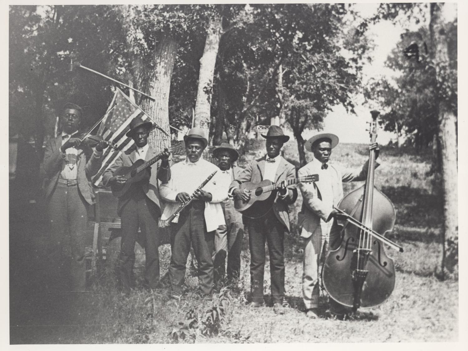 A historical photograph showing Five Black men wearing hats, jackets and ties, and holding various musical instruments, posing for an outdoors formal photograph.