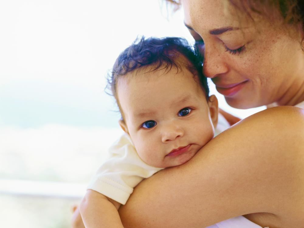 A woman holds a baby, who is looking over her shoulder into the camera. The woman is looking at the child’s head.