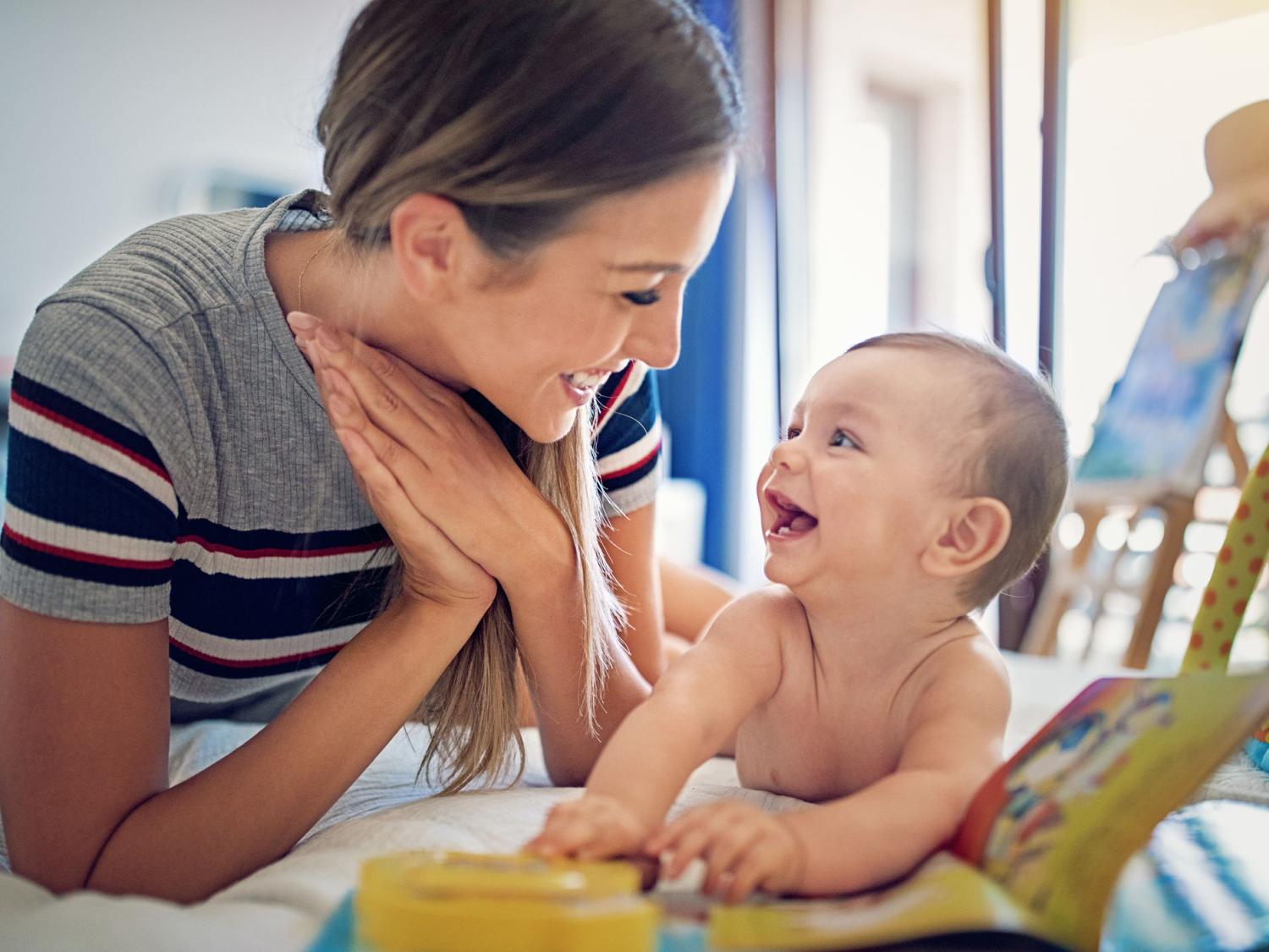 A woman and a baby lay side by side on their stomachs. They look at each other, smiling. The baby has their hands on a book and a toy in the foreground.