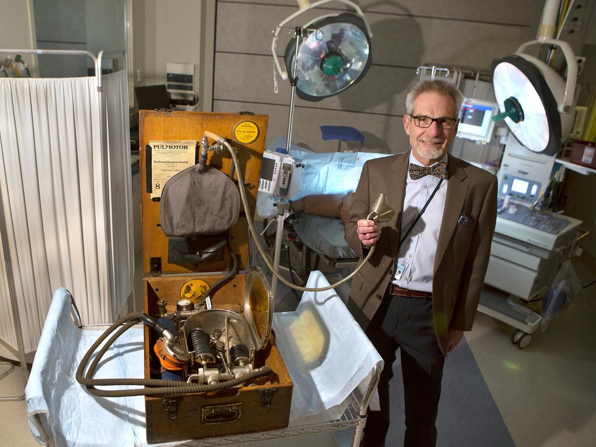 Dr. Berend Mets stands next to an old machine in a wooden case. He holds a mouthpiece at the end of a hose attached to it. He stands in a hospital room, surrounded by medical equipment.