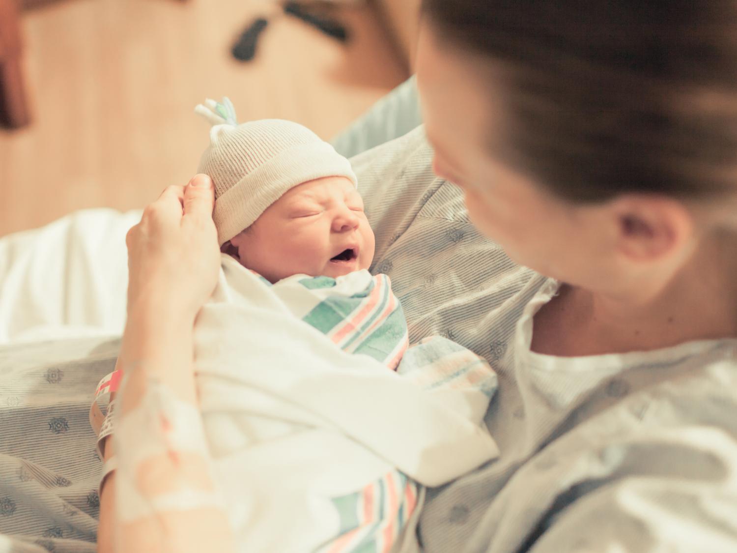 A mother holds a newborn baby swaddled in a blanket and wearing a hat