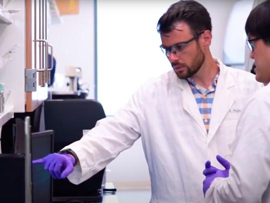 A man in a white lab coat and gloves points to a computer screen, while another man with gloves and a lab coat watches. 