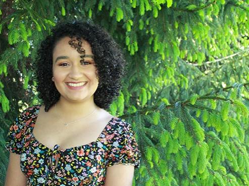 A young woman wearing a floral and paisley print top smiles at the camera in front of a green tree. 