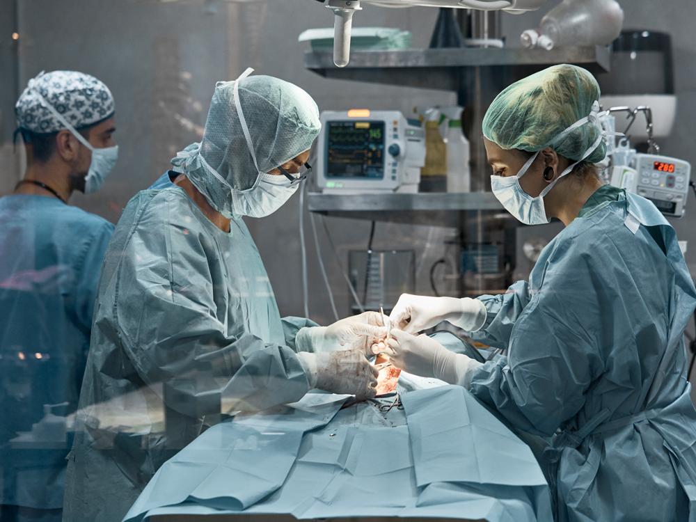 Two medical staff perform surgery on a patient. Each staff member is wearing full surgical scrubs, a mask, head covering and gloves. A large light is overhead, and various medical equipment and two other medical staffers are in the background.