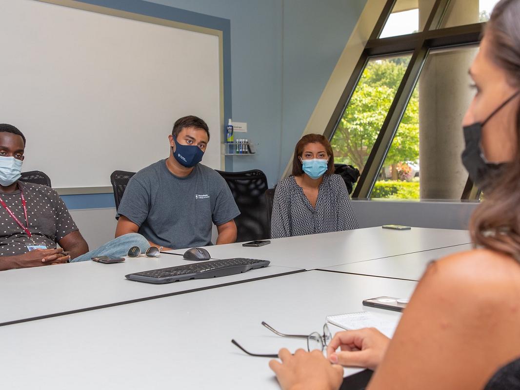 Three people in masks sit at a table speak to another woman in a mask.