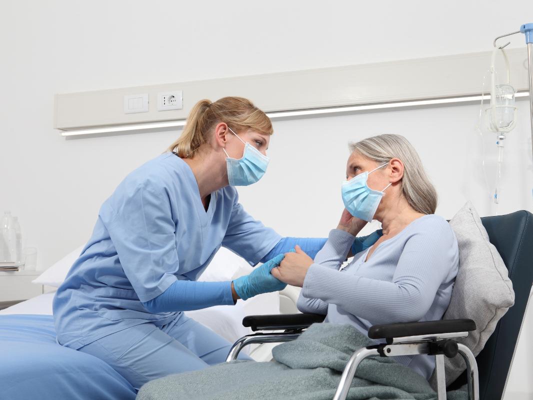 A nurse comforts an elderly woman in a wheelchair by holding her hand and placing her other hand on the patient’s shoulder.