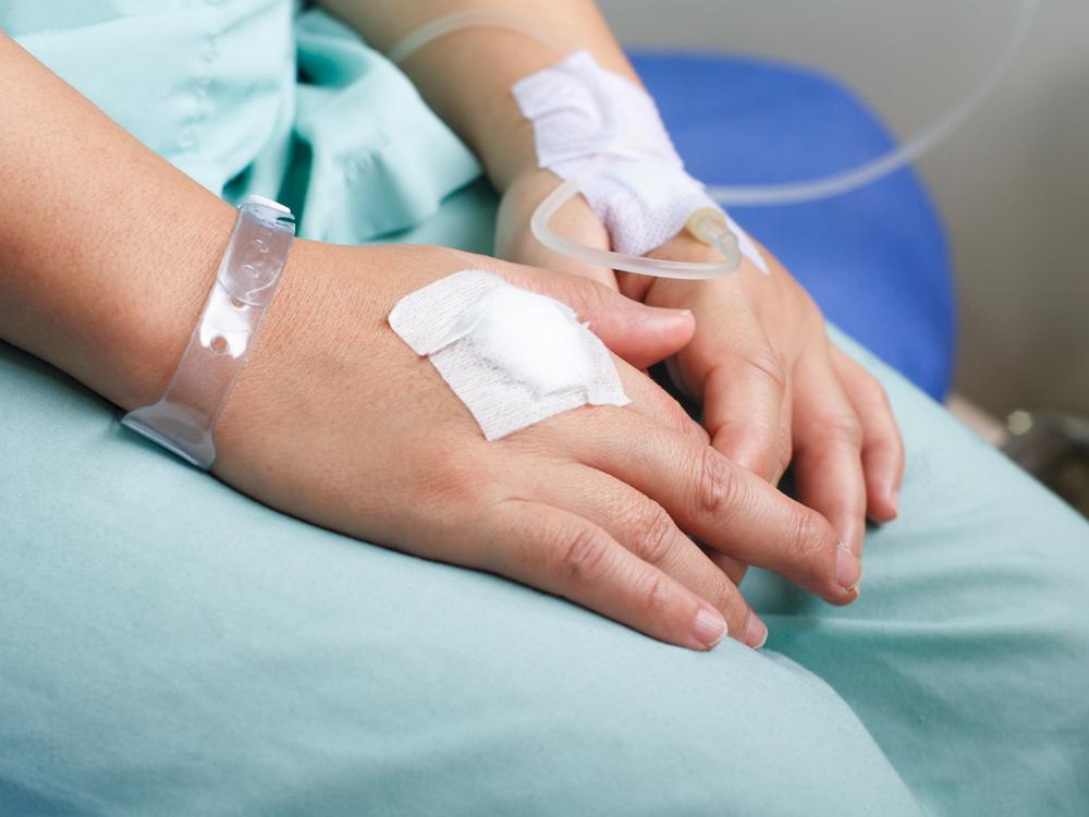 A close-up of a hospital patient’s hands, partially folded in their lap. One hand is connected to an IV line, the other has a white bandage on it.