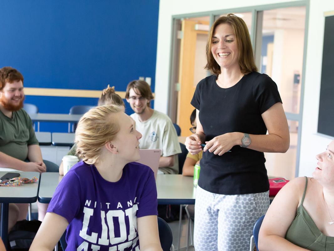 A woman in a black shirt stands smiling as she teaches a group of students who are seated around her.