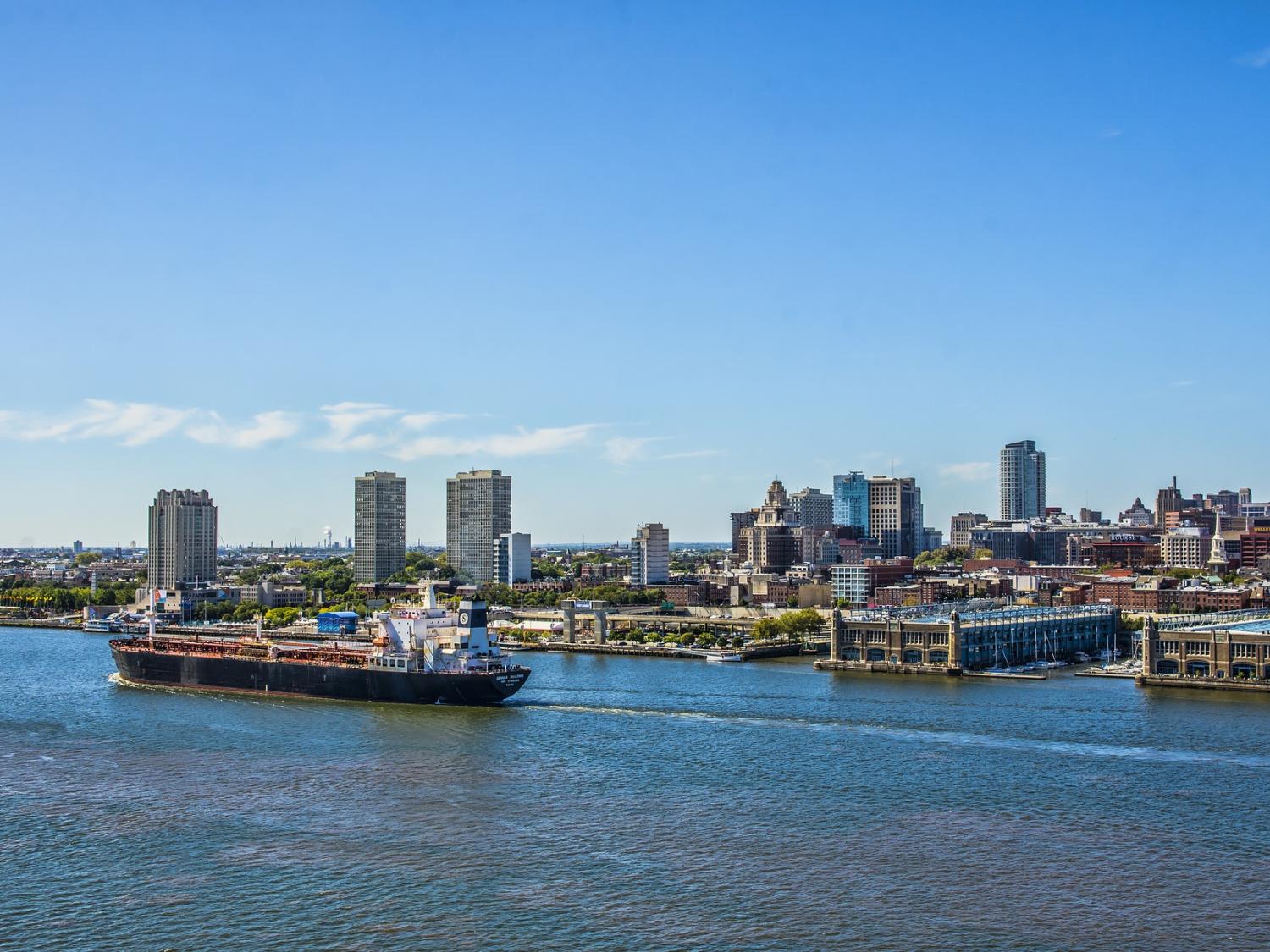 A barge passes in front of a riverfront marina with buildings and skyscrapers in the background