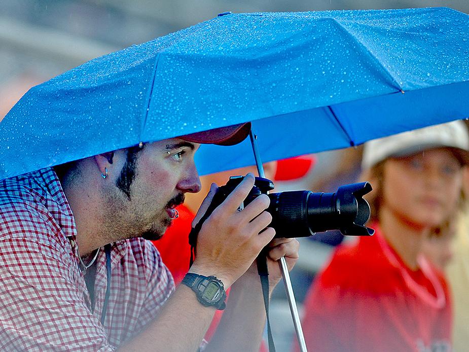 A photographer works in the rain under an umbrella to complete an assignment.