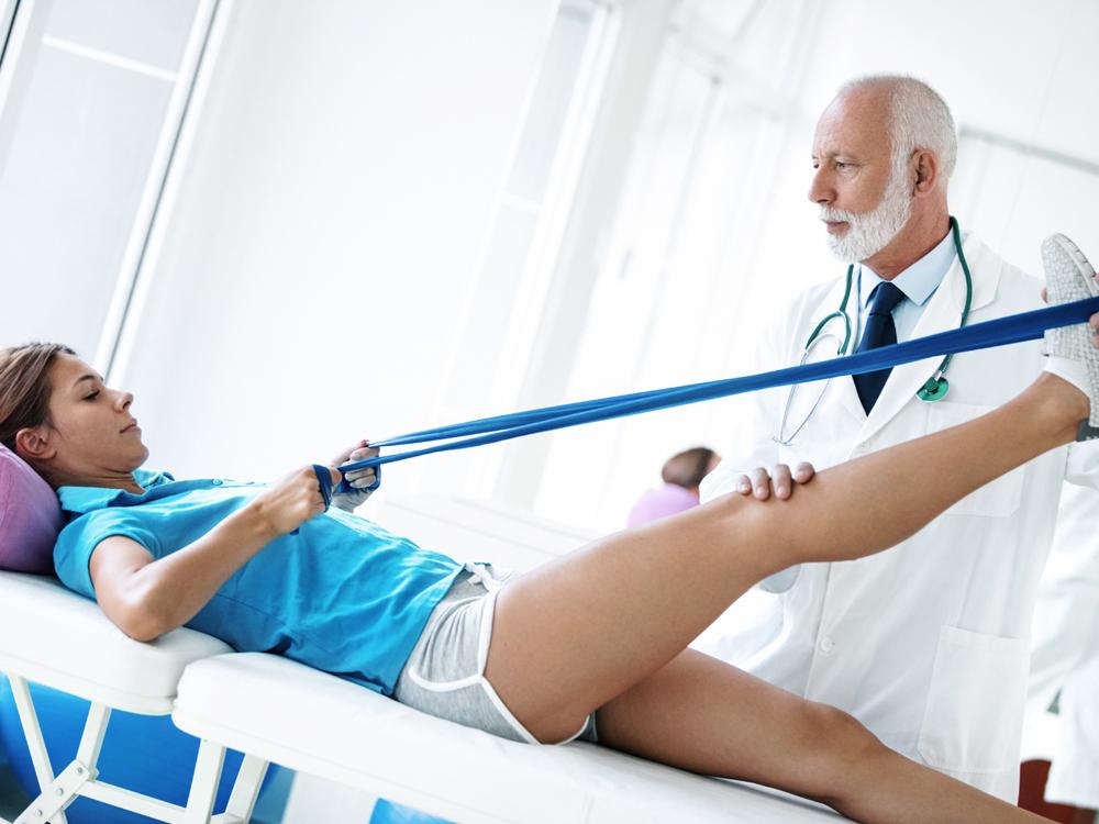 A woman lays on her back on a table, holding a band with both hands. It’s looped around her right foot, which is raised in the air. A man in a physician’s coat and stethoscope stands next to the table, holding her foot and knee.