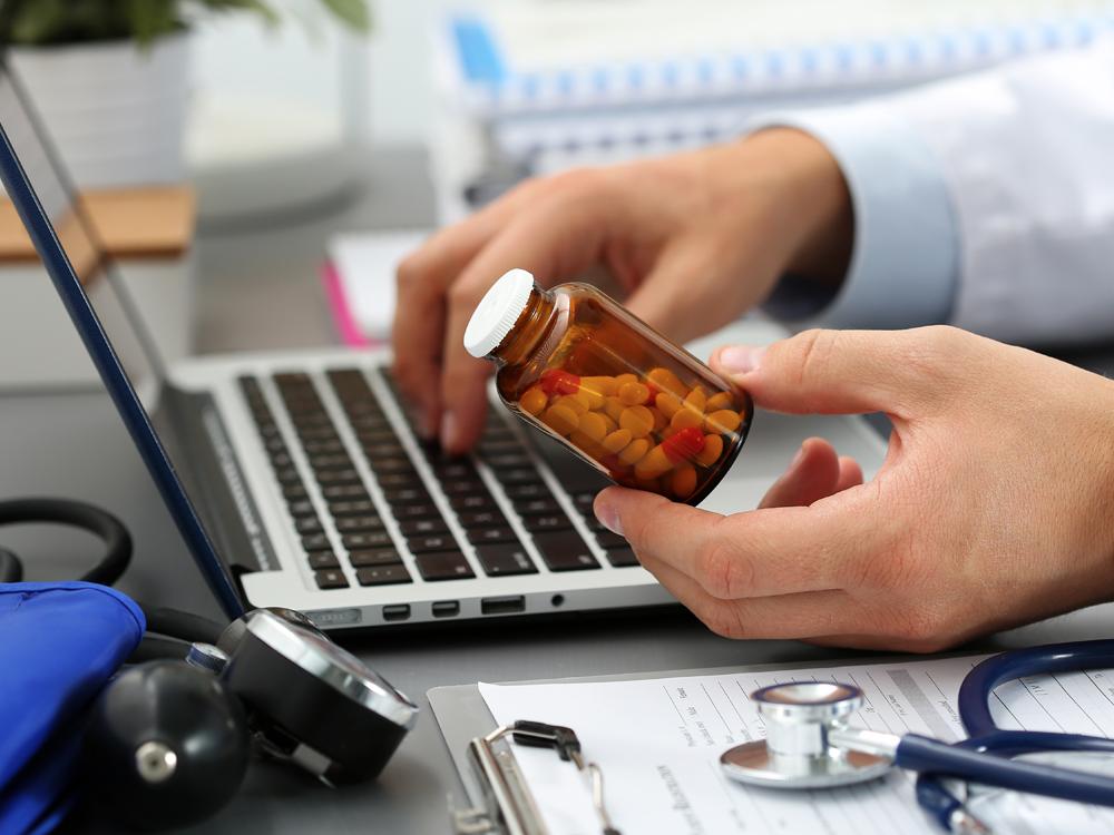 A close-up side view of a laptop computer with one hand typing on the keyboard and the hand nearest the camera holding a bottle of pills. Also on the desk is a blood pressure cuff, stethoscope and a clipboard with some papers on it.