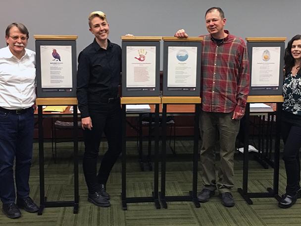 photo of two men, two women standing next to vertical poster stands holding their winning poem's poster design