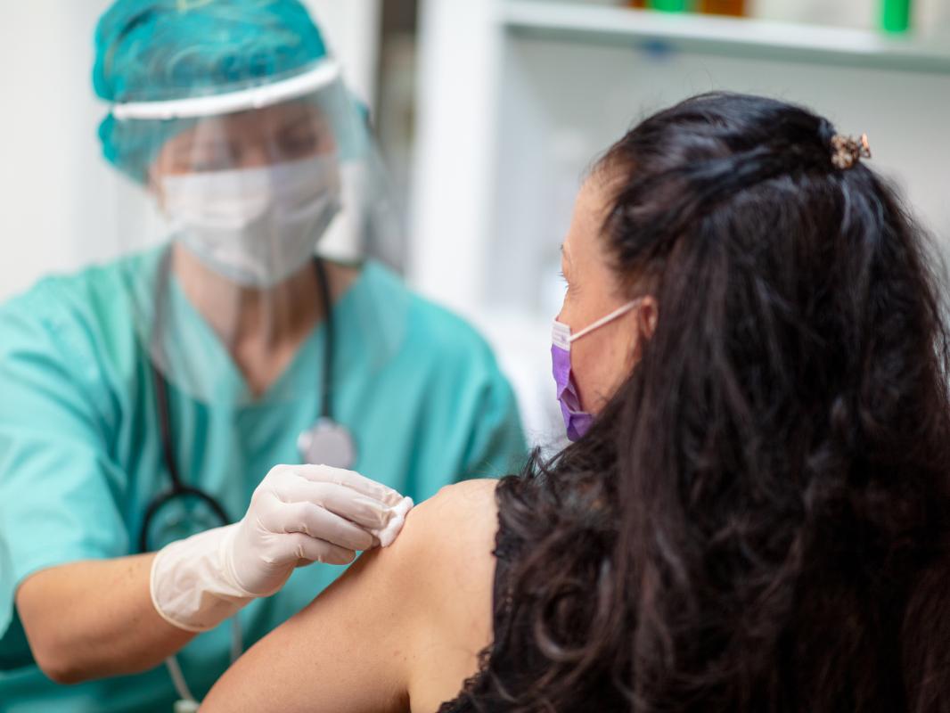 A female medical staffer wearing scrubs, a mask, a face shield and a stethoscope wipes the left arm of a female patient, who faces away from the camera.