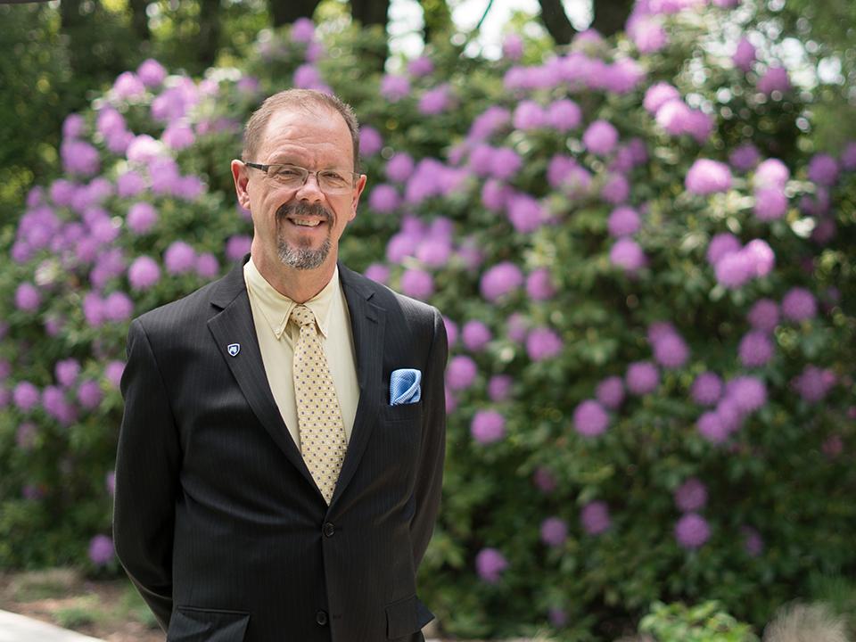 Man in a suit standing in front of a bush with purple flowers.