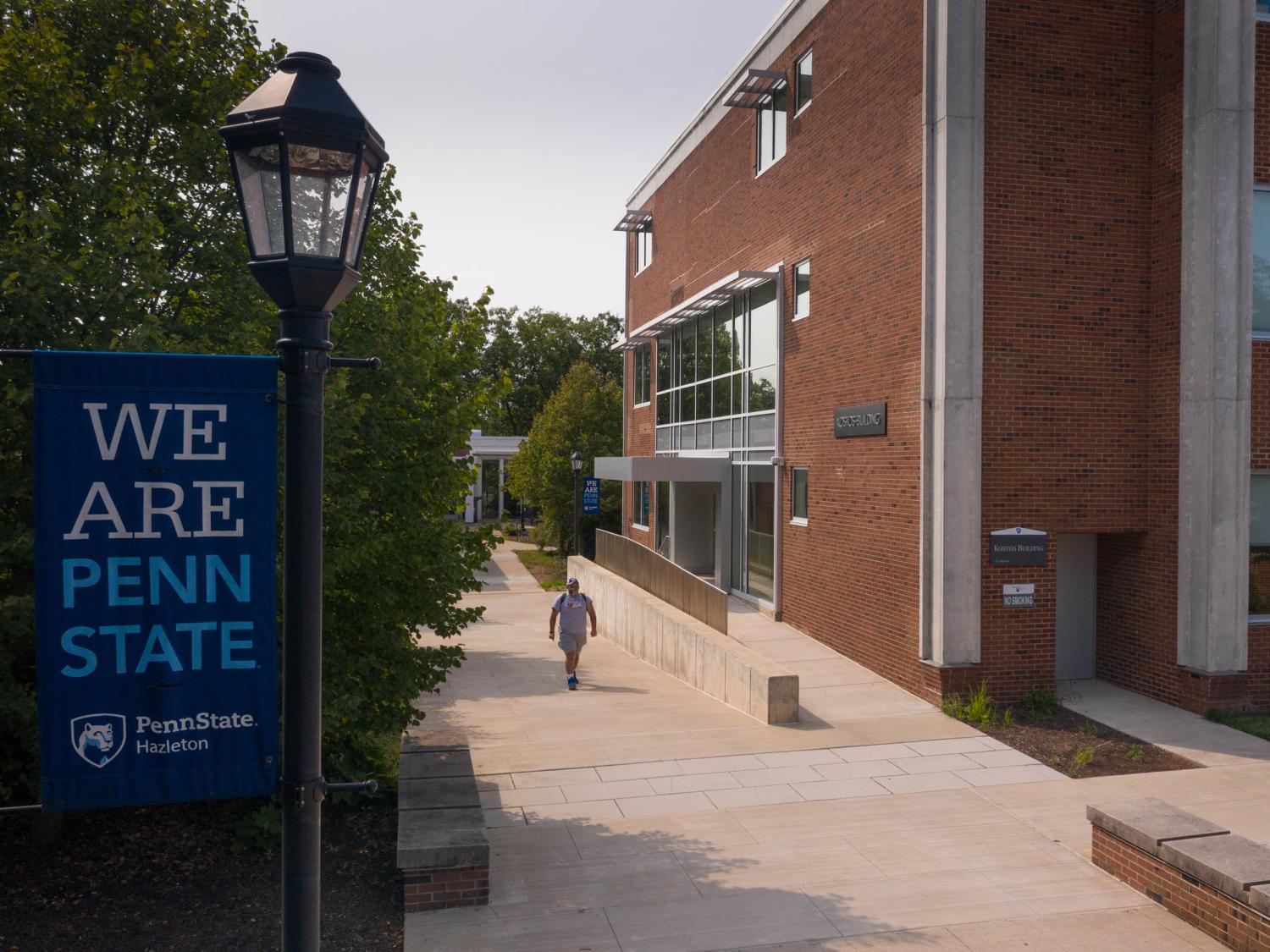 Student walking along sidewalk in between trees and a large brick building.