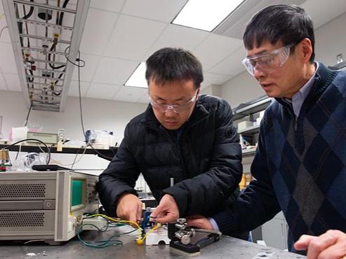 Two men wearing safety goggles adjust wires on a device in a lab.