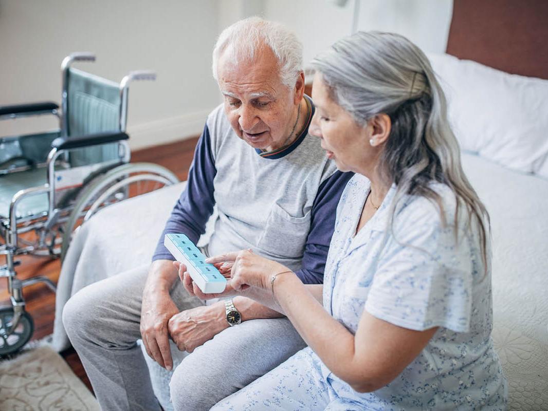 A woman sits next to a man on a bed, holding a pill container with days of the week marked on it. She points to it as she talks. A wheelchair is in the background.