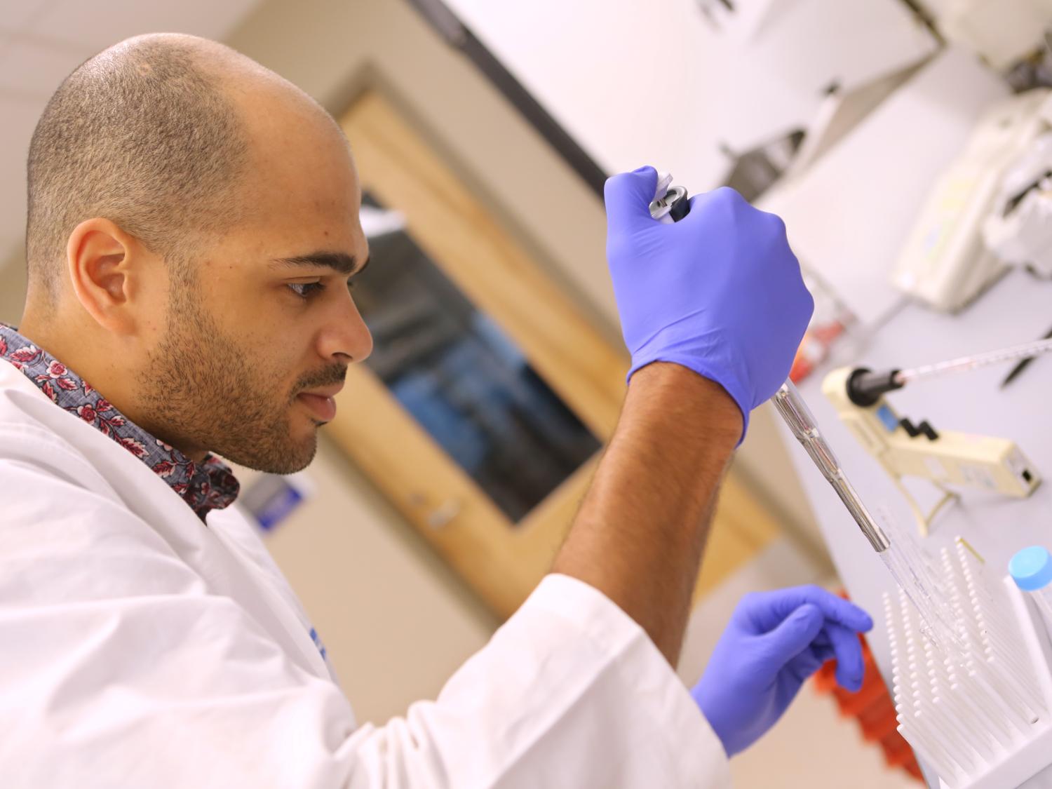 A man in a white lab coat and blue gloves uses pipetting equipment in a lab.
