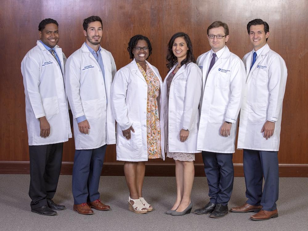 Six men and women in lab coats smile at the camera as they stand in front of a wood wall.