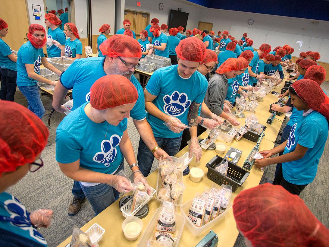 Several people stand along tables in a large room, each wearing a hairnet and working together to pour measured amounts of food into bags.