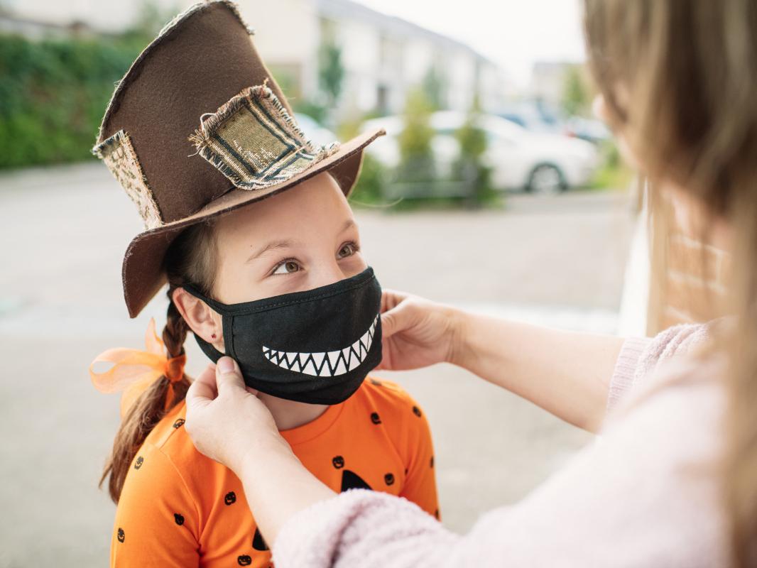Young girl dressed for Halloween in brown top hat and pumpkin shirt gets her black smiley-face cloth facemask fitted by her mother prior to trick-or-treating.